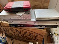 Photo shows 3 DVD players stacked on a wooden box, one Philips branded, silver and gray colors, the box has large faded letters.