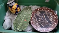 Photo showing a colorful ceramic vase, a green ceramic leaf-shaped dish, and a large round ceramic plate with tribal design inside a green tub