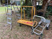 Outdoor photo showing a yellow hand truck with wooden platform alongside a metal pool ladder leaning against a tree.
