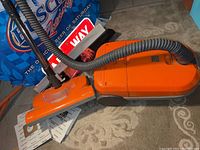 Orange Kenmore upright vacuum cleaner with grey hose and vacuum head on carpet, brand label and feature markings visible