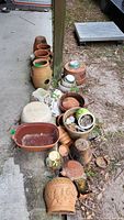 Top-down image showing a variety of assorted clay pots lined along a patio edge and ground, some stacked, in weathered, dirty condition with moss and debris.