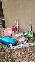 Overview of lot items grouped together outside against wall 'Green Works Pressure Washer, Rowenta Aqua control, gas cans, rolling tank, stop sign'