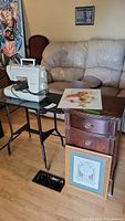 Show full setup with sewing machine on black metal stand, wooden end table with drawers, framed cross-stitch art on floor, and tin wash sign leaning against end stand.
