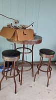 Round rusted metal table with two matching rusted metal stools that have wooden tops. The table and stools show heavy weathering and rust. Items are shown indoors on a concrete floor.