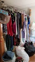 Wide view of assorted women's dresses, scarves, and other clothing items on a wooden rack, with bags filled with clothing and shoes on the floor.