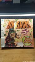 Front of game box showing title Dinosaurs Gotta Love Me! with cartoon dinosaur characters and branding as Milton Bradley.