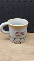 Clear front view of Krispy Kreme mug showing the logo, yellow rim and green polka dot border on white ceramic.