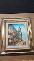 Front view of the oil painting showing a street scene with a tower, framed in a gold-tone multi-layered frame.