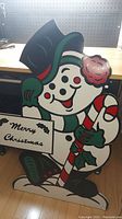 Front view of the wooden snowman holding a candy cane and Merry Christmas sign indoors on wooden floor.