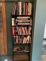 Full view of metal framed shelf filled with various books, bookshelf against green wall with wooden panel on left.