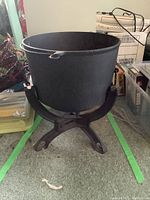 Side view of black cast iron cauldron on matching cast iron stand with three legs and holes for securing base.
