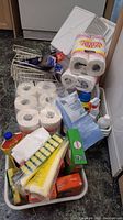 Lot showing multiple packs of white paper towels, plastic baskets filled with cleaning liquids, sponges, aluminum foil, and paper product boxes.