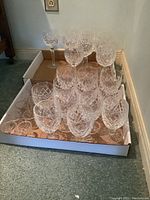 Set of 12 water glasses and 9 wine glasses shown inside a shallow white cardboard tray on carpet floor near baseboard and electrical outlet.