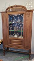 Front view showing the carved solid walnut china cabinet with decorative iron scrollwork on glass door and three interior shelves with items inside.