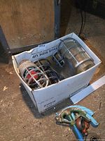 Box showing three metal industrial light cages and old brass propeller on floor, cage glass covers visible on two of them