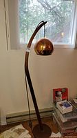 Full lamp showing round teak base, curved arm, hanging copper globe shade, black cord and footswitch.
