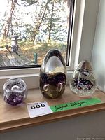 Three signed art glass paperweights displayed in a row on a wooden surface by a window with outdoor view. Each is distinct in shape and internal design.