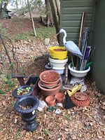 View of entire lot showing cast iron planters, clay pots, resin planters, plastic heron, concrete frogs, wooden planter cart and garden items on ground covered with leaves.