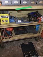 Shelf with various tools including electric drills, a yellow boxed item, metal brackets, and tool cases beneath.