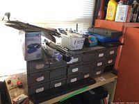 Metal 17-drawer cabinet with drawers labeled and one missing, containing various small items and tools inside. On top are multiple hand tools, boxes, and containers.