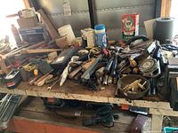 Wide view of a cluttered workbench filled with various hand tools including saws, hammers, screwdrivers, levels, and other workshop tools.