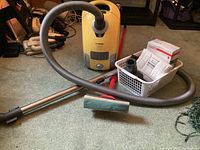 Photo showing yellow Miele canister vacuum cleaner with hose and metal wand laid out on carpet, including a sponge mop in front