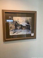 Framed watercolour painting showing snowy cabin and trees from a distance hung on a wall.