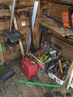 Overall photo showing red fuel can, hand tools, baskets with smaller tools, pick axe and hatchet leaning against the wall, and yellow trouble light and booster cables on shelf.