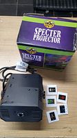 Spirit Halloween Specter Projector, black unit with ventilation slots and power cord, alongside box and film slides