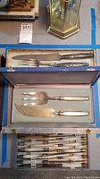 Top view of a box with a large serving knife and fork with ornate silver handles, and additional cutlery with white handles below in separate compartments.