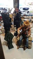View of four main items: two black carved Asian statues, green dragon statue, and cowboy animatronic figurine sitting and playing violin on tabletop with other plates and decor in background.