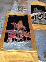 Three silk Asian tapestries laid out showing detailed embroidery of figures, horses, dragon, and bridge with golden yellow borders on black fabric.