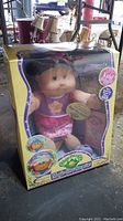 Full view of the doll in its original yellow box packaging with clear front window, showing the doll inside and branding.