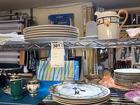 Metal shelf displaying a stack of white plates and a ceramic pitcher with painted fruit design, surrounded by porcelain items on a worktable
