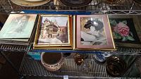 Photo showing four framed art prints including a vintage architecture print, classical portrait, floral print and more on metal shelving