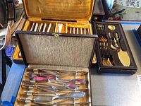 Two sets of cutlery arranged on a table; one silver-toned set in a beige case with multiple butter knives and forks, and a gold-plated set in a yellow-lined box with various forks and spoons.
