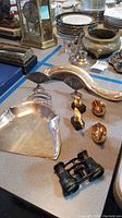 Photo showing antique silver dustpan along with the vintage opera glasses and three gold-tone swan figurines on a table with other items in background.