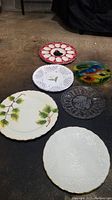 Full view of a variety of decorative and serving plates on dark floor including Lenox platter and other artistic glass and ceramic plates.
