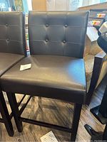 Close-up of one black bar stool showing tufted backrest and wooden legs on wood floor.