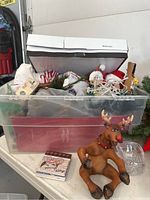 Clear plastic bin filled with assorted Christmas decorations including string lights, small ornaments, and a brown plush reindeer figurine in front.