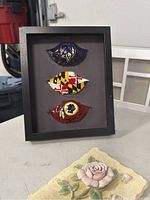 Black wooden framed shadowbox holding three glass Baltimore Ravens art pieces shaped like eyes in different color patterns.