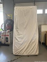 Front view of hospital bed mattress covered in fitted white cover standing upright.