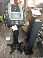 Full view of karaoke machine on stand with built-in screen and control panel. Two tall black external speakers standing next to it. Wired microphone attached to the machine. Cables around the base.