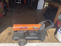 Orange and black ALL+PC 170T kerosene heater with wheels and handle placed on a wooden floor in a garage or workshop setting with visible signage and tools in background.
