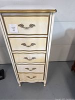 Front view of the wooden lingerie chest showing cream color and five drawers with ornate gold-tone handles.