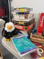 Photo of stacked jigsaw puzzle boxes (375, 500, 1000 pieces), plush eagle, vinyl pillow cover, and small wicker basket on a table.