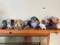 Wide shot showing collection of stained glass figurines and decor on wooden mantle, including butterflies, clown, heart frame, and flowers.