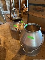 Two vintage metal milk cans placed on a dirt floor showing the shape, handle, and opening of each can.
