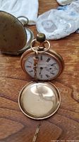 Gold-tone hunter-case pocket watch with front cover open showing white enamel dial with Roman numerals and subsidiary seconds; second watch case visible in background