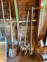 Photo showing all four tools leaning against wooden wall: shovel, pickaxe, post hole digger, tamper.